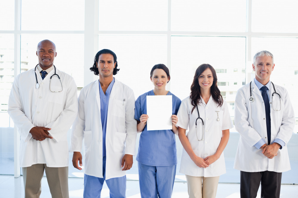 Four individuals, likely doctors or nurses, stand in a row against a bright, light-filled background of large windows.
